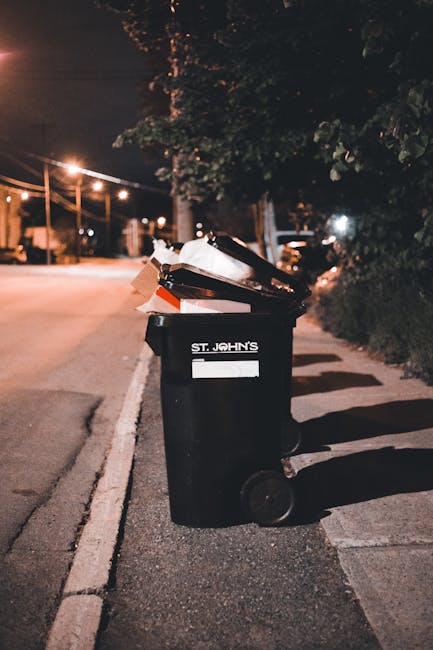 A black wheeled rubbish bin labeled with the name 'St. John's' is positioned on a quiet urban street at night, with streetlights casting a warm glow. The bin is filled with various waste items, including cardboard boxes, paper bags, and plastic packaging, some of which are protruding from the top. The bin is situated on the paved pavement near the edge of the street, adjacent to a curb that separates it from the asphalt road. In the background, there are trees with dark foliage, and blurred streetlights and parked vehicles, indicating a residential or suburban area. The scene is illuminated by artificial lighting, creating soft shadows on the ground and a slightly subdued atmosphere. This image exemplifies the type of typical waste or rubbish that might be queued for collection or removal, aligning with the waste management services provided by Waste Removal Tufnell Park that support private disposal or alternate rubbish collection methods outside of local authority services.