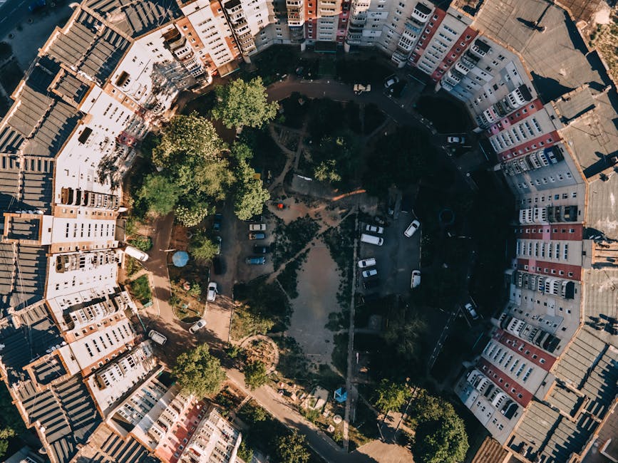 An aerial view of a circular residential courtyard surrounded by multi-storey terraced buildings with white, beige, and red brick facades. The courtyard features a central green area with several mature trees, pathways, and patches of grass. Multiple parked cars are visible along the perimeters and within designated parking spaces, with some vehicles parked parallel to the buildings and others angled. The surrounding buildings have pitched roofs with gray tiles and numerous windows, creating a dense urban environment. Shade and sunlight interplay across the scene, casting shadows from the trees and buildings. Overall, the image captures a quiet, inhabited residential complex, illustrating an environment where private waste collection services like those of Waste Removal Tufnell Park may operate for rubbish removal or site clearance within the courtyard area. The scene emphasizes the compact layout typical of inner-city residential estates, with open spaces for communal use, underlying the importance of regular rubbish collection and waste management for local residents.