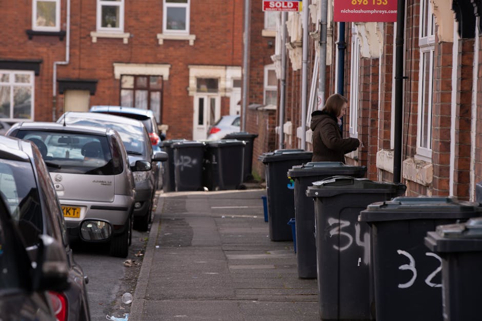 A black wheeled rubbish bin labeled with the name 'St. John's' is positioned on a quiet urban street at night, with streetlights casting a warm glow. The bin is filled with various waste items, including cardboard boxes, paper bags, and plastic packaging, some of which are protruding from the top. The bin is situated on the paved pavement near the edge of the street, adjacent to a curb that separates it from the asphalt road. In the background, there are trees with dark foliage, and blurred streetlights and parked vehicles, indicating a residential or suburban area. The scene is illuminated by artificial lighting, creating soft shadows on the ground and a slightly subdued atmosphere. This image exemplifies the type of typical waste or rubbish that might be queued for collection or removal, aligning with the waste management services provided by Waste Removal Tufnell Park that support private disposal or alternate rubbish collection methods outside of local authority services.