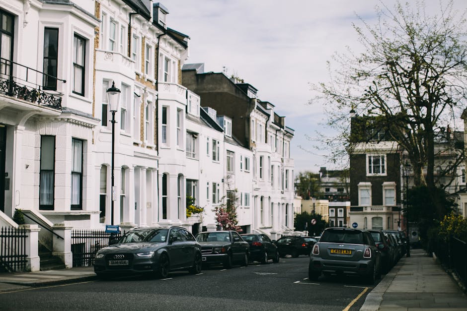 The image depicts a residential street lined with white Victorian-style terraced houses, featuring ornate architectural details and bay windows. Several cars, including an Audi, a Mini, and other vehicles, are parked along the pavement in front of the houses. The street is bordered by a black metal fence and a sidewalk, with a few trees visible, including one large tree with sparse branches and small leaves on the right side. The sky appears overcast, casting diffuse natural light across the scene. The area looks quiet and well-maintained, characteristic of urban neighbourhoods where private waste collection or rubbish removal services, such as those by Waste Removal Tufnell Park, are often employed for dealing with household waste outside of municipal rubbish collection, especially during house clearance projects or property renovations.