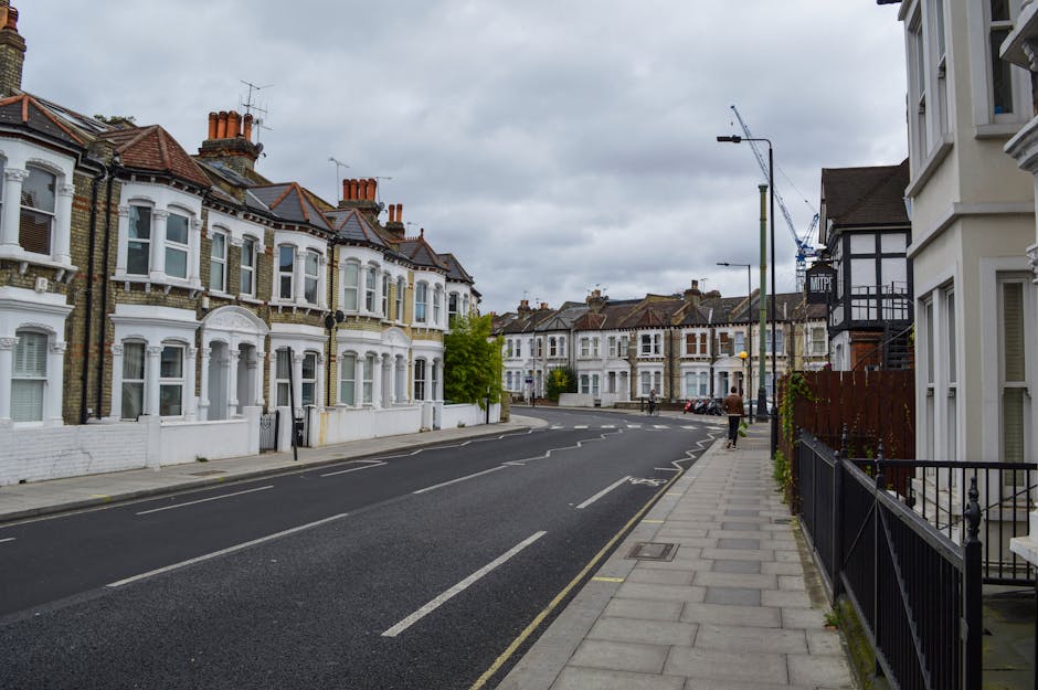 A residential street in a suburban area with Victorian-style terraced houses on the left side, featuring brick facades with white-painted details and bay windows, and some houses having small front gardens enclosed by low fences. The street appears to be relatively quiet, with a few parked cars along the curb and a solitary pedestrian walking on the paved sidewalk on the right side, which is lined with black iron railings. The road has clearly marked parking and driving lanes, with a gentle curve into the distance under overcast, gray skies that cast diffuse lighting over the scene. Streetlights and a construction crane are visible in the background, indicating ongoing development in the area. The overall environment appears well-maintained and typical of suburban housing areas, fitting within the context of private waste management and alternative rubbish collection practices supported by Waste Removal Tufnell Park, as suggested by the surrounding urban setting.