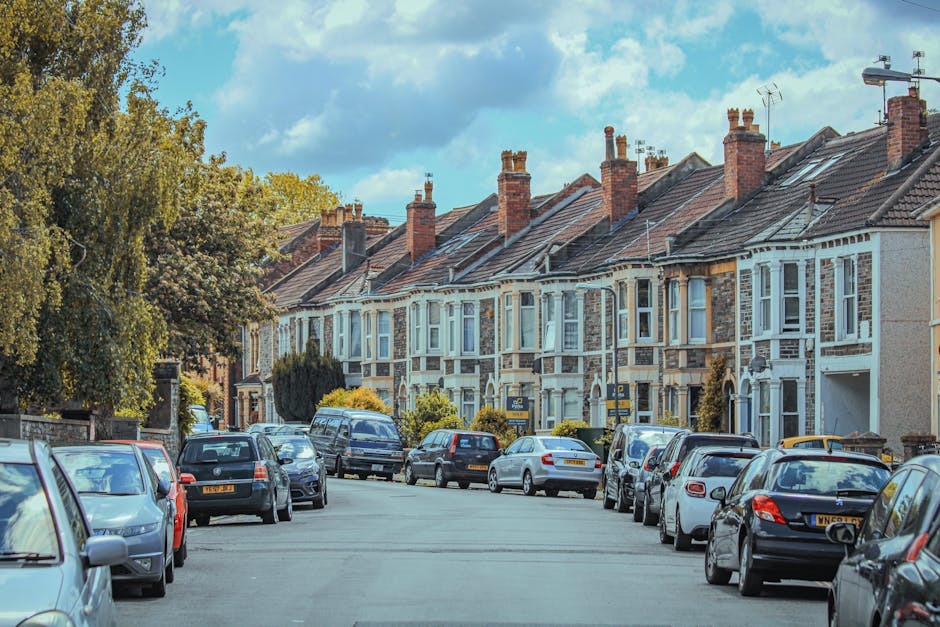 The image depicts a residential street lined with white Victorian-style terraced houses, featuring ornate architectural details and bay windows. Several cars, including an Audi, a Mini, and other vehicles, are parked along the pavement in front of the houses. The street is bordered by a black metal fence and a sidewalk, with a few trees visible, including one large tree with sparse branches and small leaves on the right side. The sky appears overcast, casting diffuse natural light across the scene. The area looks quiet and well-maintained, characteristic of urban neighbourhoods where private waste collection or rubbish removal services, such as those by Waste Removal Tufnell Park, are often employed for dealing with household waste outside of municipal rubbish collection, especially during house clearance projects or property renovations.