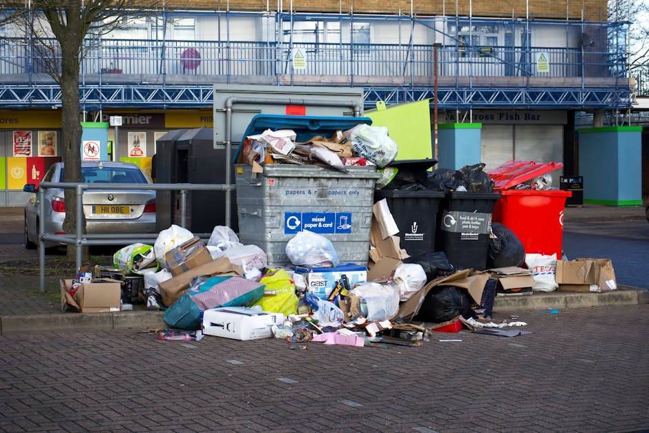 A large accumulation of mixed rubbish and waste materials is piled against a metal barrier on a paved sidewalk in an urban setting during daylight. The debris includes various discarded cardboard boxes, paper bags, plastic packaging, and black trash bags, some of which are torn or spilling contents. A prominent grey waste bin labeled for mixed paper and cardboard is overflowing, with additional waste placed around it, contributing to the cluttered appearance. Behind the waste are parked cars, one grey and partially visible, and a row of brightly coloured storefronts with signage, indicating a commercial area. In the background, there is a building under construction or refurbishment, covered with scaffolding and safety netting. The scene reflects a typical case of unmanaged refuse that may be handled by private rubbish removal services like Waste Removal Tufnell Park, often used for on-site clearance or alternative waste handling outside municipal collection schedules.