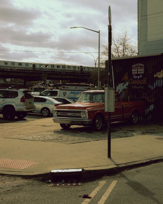 The image depicts a daytime urban scene with a vintage red pickup truck parked beside a small building with signage indicating it is a retail or garage space, positioned on a concrete pavement. The truck has a metallic finish, a classic boxy design, and appears to be in good condition. Nearby, several modern vehicles including a white SUV and a silver sedan are parked along the street, with a police van visible further back. An elevated train is passing in the background on a rail track supported by concrete pillars, with grey clouds overhead contributing to diffused natural lighting. A streetlight extends from a pole next to the truck, which is situated on a sidewalk that has tactile paving for pedestrian safety, and a drainage grate is visible at the curb. There is a leafless tree behind the scene, indicating a possible winter or early spring season. The environment suggests a typical city street where private vehicle parking and urban infrastructure are present, aligning with the context of alternative waste handling and rubbish disposal support services provided by Waste Removal Tufnell Park.