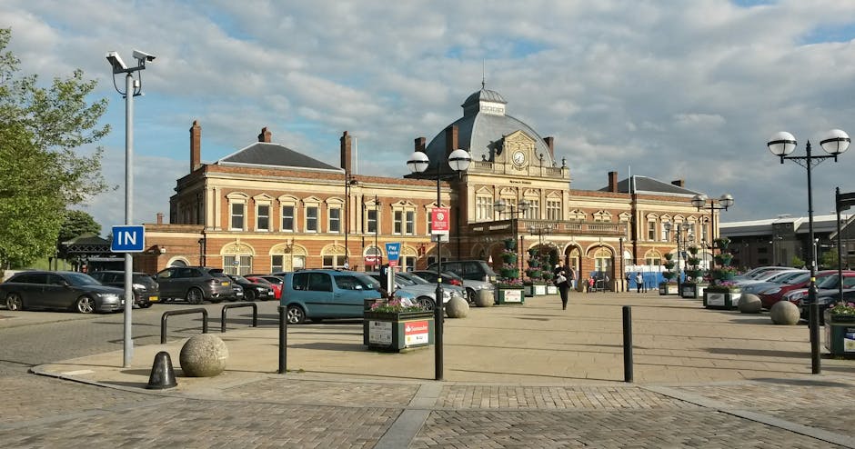 A large historic railway station building constructed in red brick and cream-colored stone, featuring arched windows and decorative architectural details, with a central tower topped by a clock and flagpole. In front of the station, the paved forecourt contains multiple parked cars, including sedans and hatchbacks, aligning along the edge of the pedestrian area. Several street lamps with dual globe lights and decorative bases are visible, along with flower planters filled with colorful blooms, placed near open metal barriers and spherical concrete bollards that separate the parking area from the pedestrian zone. The environment appears to be in daylight, with a partly cloudy sky overhead, and a few people walking across the forecourt. The scene represents an urban setting facilitating independent or private waste collection, fitting within the context of rubbish removal services offered in the area, such as those by Waste Removal Tufnell Park.